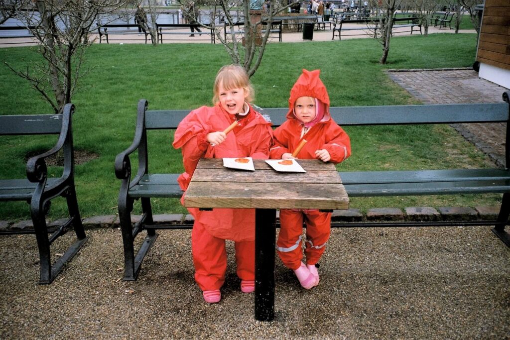 To piger i 6 års alderen spiser pølser på bænk i rødt regntøj