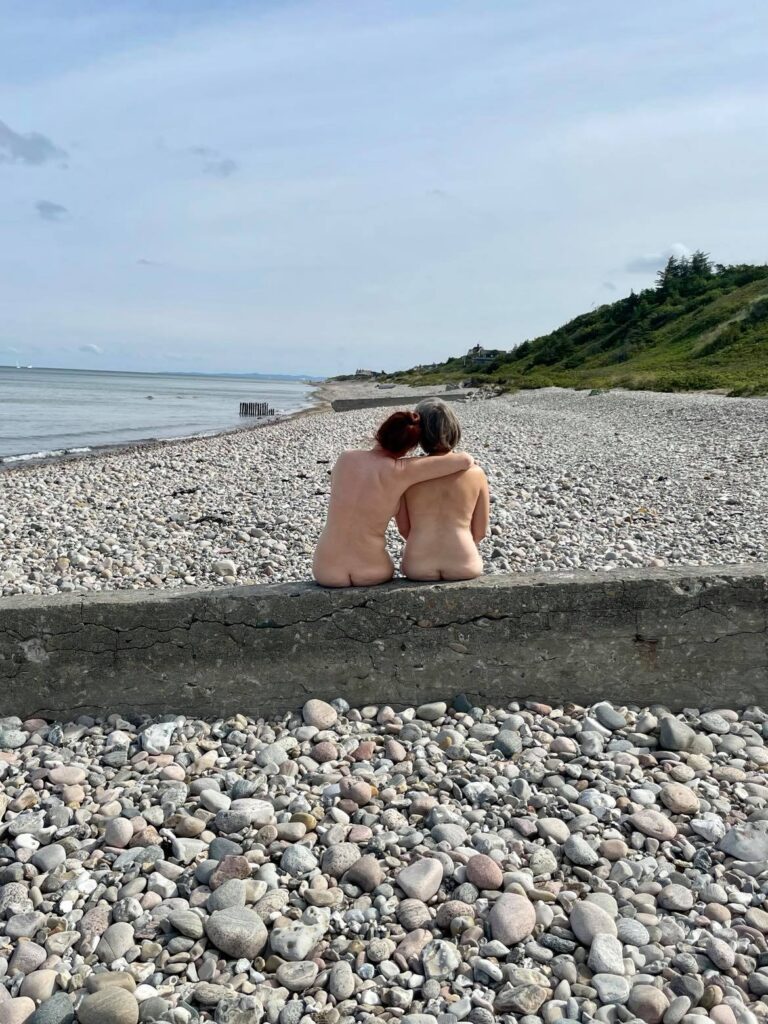 To kvinder uden tøj set bagfra siddende på en mur på en strand